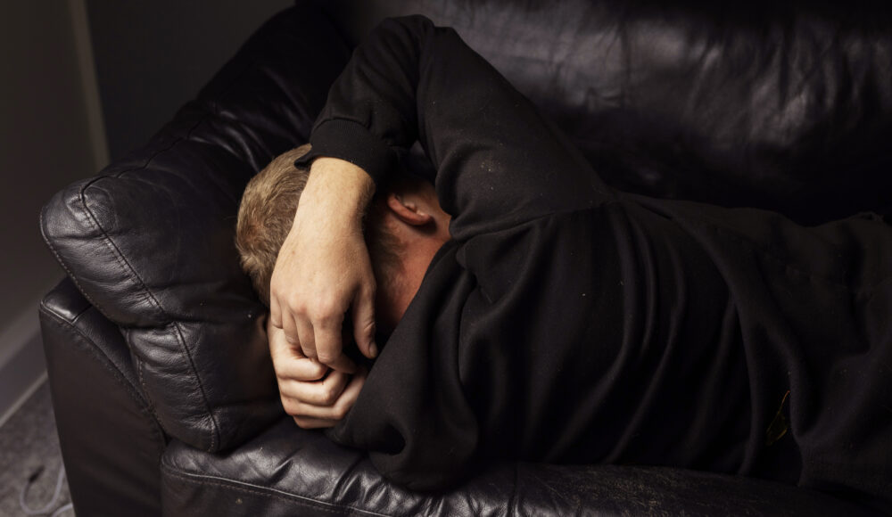 En person ligger sammenkrøbet på en mørk lædersofa med armen over hovedet i et dæmpet rum.