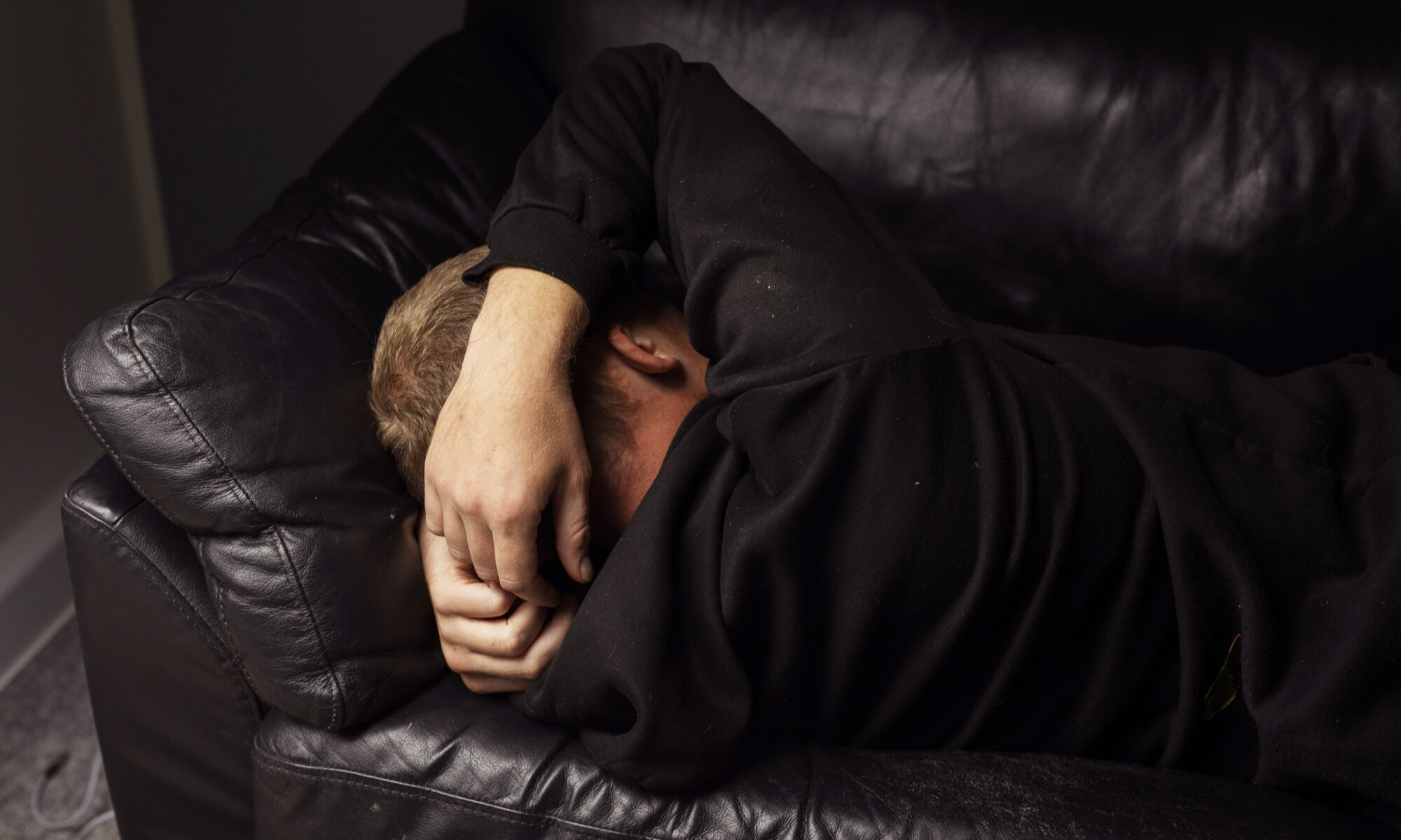 En person ligger sammenkrøbet på en mørk lædersofa med armen over hovedet i et dæmpet rum.