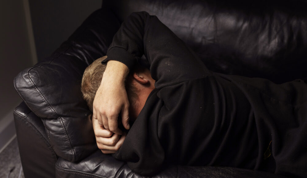 En person ligger sammenkrøbet på en mørk lædersofa med armen over hovedet i et dæmpet rum.